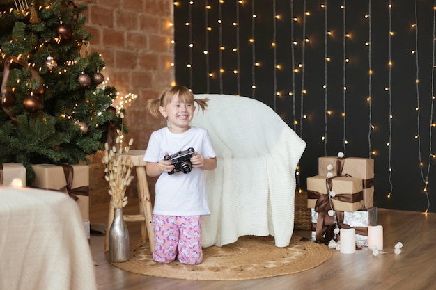 Adorable petite fille de 4 ans en pyjama est assise sur le sol avec un appareil photo dans ses mains près de l'arbre de Noël.