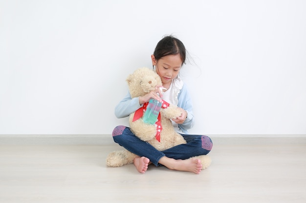 Adorable petite fille asiatique enfant appliquant un gel d'alcool à partir d'une bouteille en plastique pour nettoyer l'ours en peluche