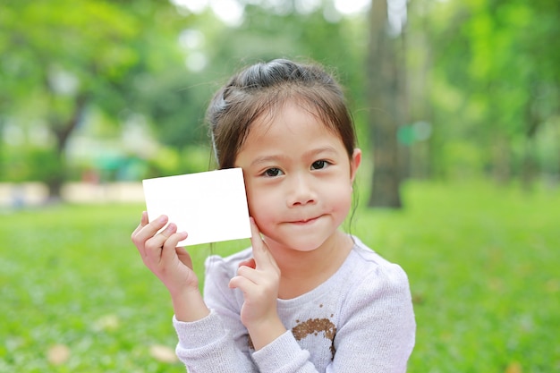 Adorable petite fille asiatique enfant montrant un papier blanc vierge dans le jardin vert.