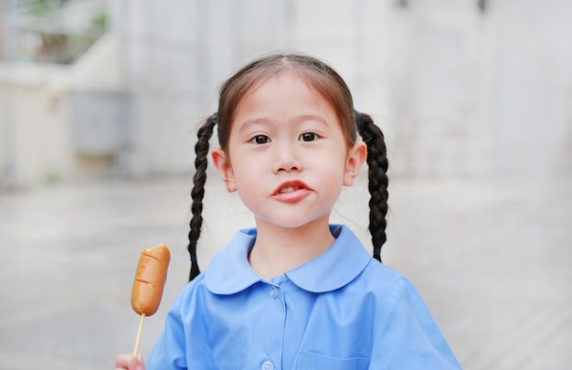 Adorable petite fille asiatique en uniforme scolaire profiter de manger des saucisses.