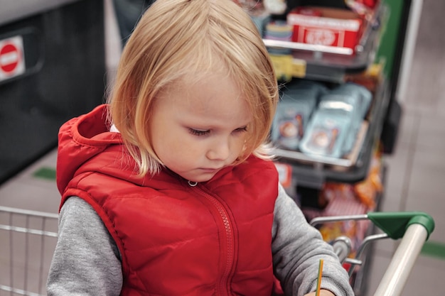 Adorable petite fille assise dans un panier en magasin ou en supermarché. Portrait petit enfant mignon faire du shopping et acheter des marchandises sur une étagère de magasin