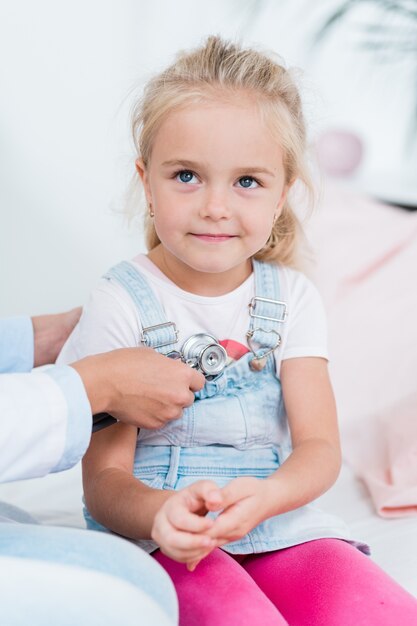 Adorable petite fille aux cheveux blonds assis sur le lit dans un cabinet médical pendant que le médecin l'examinait avec un stéthoscope