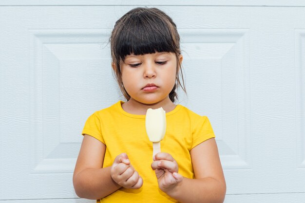 Adorable petite fille aux cheveux noirs, manger de la crème glacée, vêtue d'une chemise jaune appuyée sur fond blanc