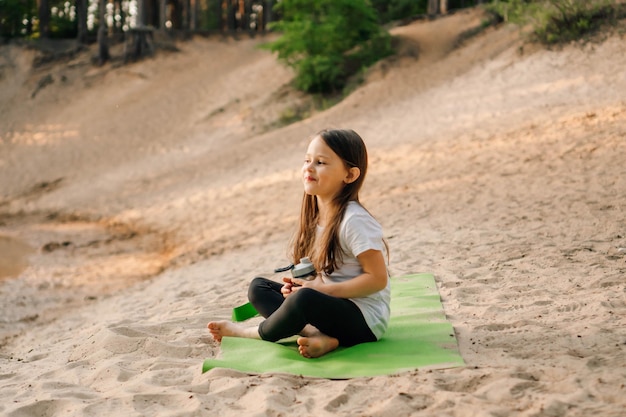 Une adorable petite fille aux longs cheveux bruns assise sur un tapis vert sur une plage de sable avec une bouteille d'eau à la main et souriante