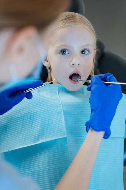 Photo adorable petite fille ayant ses dents vérifiées par une femme dentiste agréable à l'aide d'un miroir buccal et d'une sonde dentaire