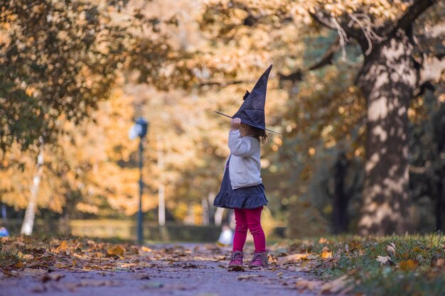 adorable petite fille en bas âge marchant dans le parc en automne. fille en costume de sorcière et chapeau noir cosplay costume d'halloween. fête d'halloween