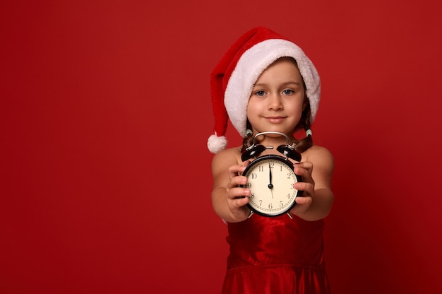 Adorable petite fille en bonnet de Noel et costume montre minuit sur l'horloge du réveil, sourit en regardant la caméra et pose sur fond rouge avec espace de copie. Bonne année, joyeux Noël