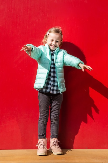 Adorable petite fille caucasienne assise sur une terrasse en bois à côté d'un mur rouge en regardant la caméra