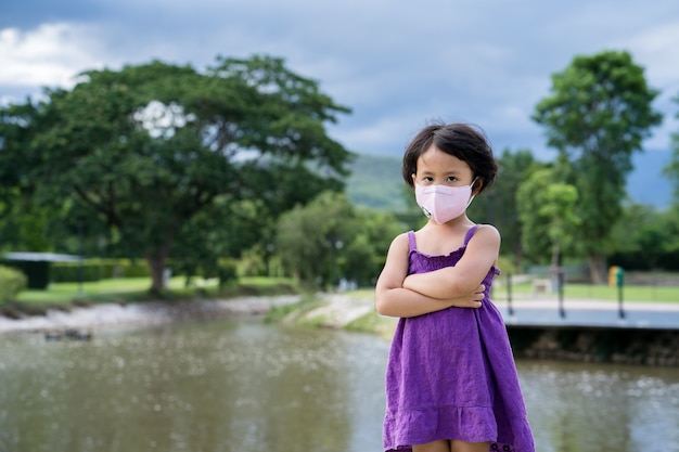 Adorable petite fille debout avec les bras croisés au parc portant un masque protecteur