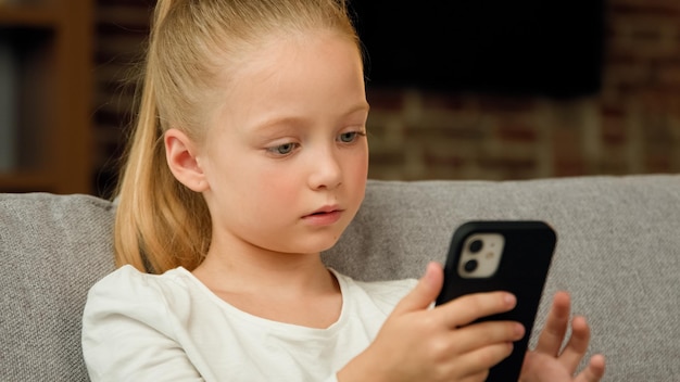 Adorable petite fille enfant mignon assis sur un canapé confortable seul dans le salon en regardant le gadget