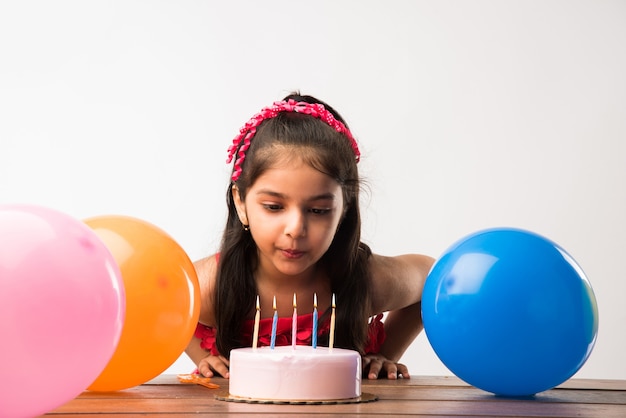 Adorable petite fille indienne ou asiatique mignonne célébrant son anniversaire tout en tenant un gâteau aux fraises et en soufflant des bougies à table ou debout isolé sur fond blanc ou rouge