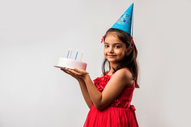 Adorable petite fille indienne ou asiatique mignonne célébrant son anniversaire tout en tenant un gâteau aux fraises et en soufflant des bougies à table ou debout isolé sur fond blanc ou rouge