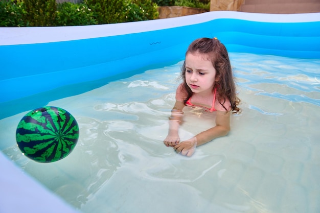 Adorable petite fille joue avec un ballon flottant tout en nageant dans la piscine gonflable par une chaude journée ensoleillée d'été