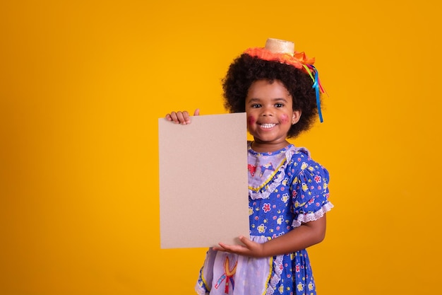 Adorable petite fille noire vêtue d'une tenue festa junina tenant une carte vierge avec espace de copie Promotion de la fête de juin