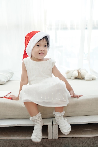 Adorable petite fille en robe blanche et bonnet de noel assis sur le lit et détournant les yeux