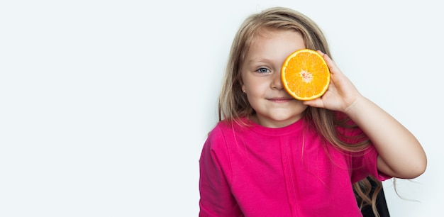 Adorable petite fille se couvre les yeux avec une orange souriant à l'avant sur un mur blanc avec espace libre