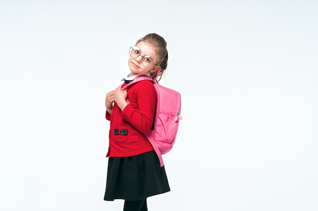 Adorable petite fille en veste d'école rouge, robe noire, lunettes arrondies tenant aux sangles d'un sac à dos et souriant et regardant la caméra, posant sur un espace blanc. Isoler