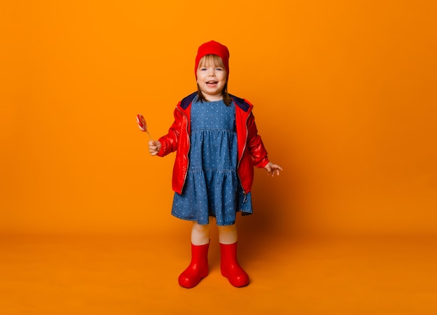 Adorable petite fille vêtue d'une veste rouge et de bottes tenant une sucette sur fond jaune. notion d'automne.