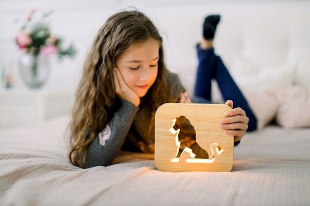 Adorable petite jolie fille allongée sur le lit dans une pièce lumineuse confortable et jouant avec une lampe de nuit en bois avec une image de lion découpée.