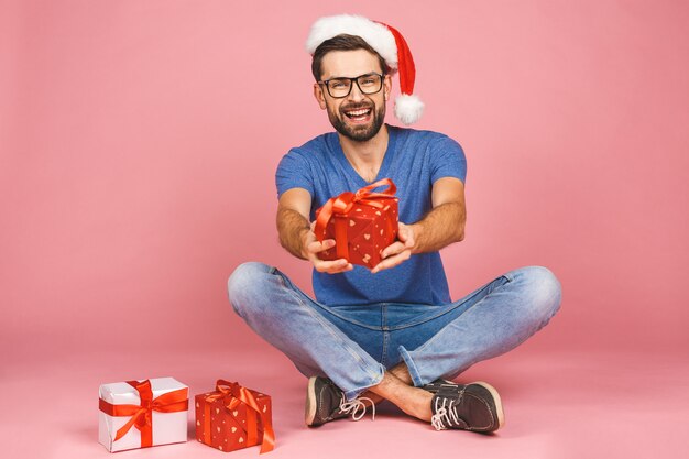 Adorable photo de séduisant jeune homme au chapeau de Noël avec un beau sourire tenant des boîtes de cadeau d'anniversaire isolées sur un mur rose, assis sur le sol. Concept de cadeau.