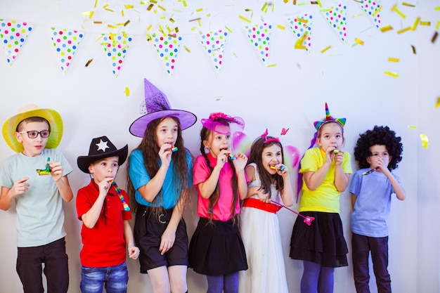 Adorables enfants joyeux s'amusent ensemble en soufflant des cornes de fête Enfants portant des chapeaux et des perruques fantaisie devant un mur décoré félicitant la fille d'anniversaire Concept enfants et événements