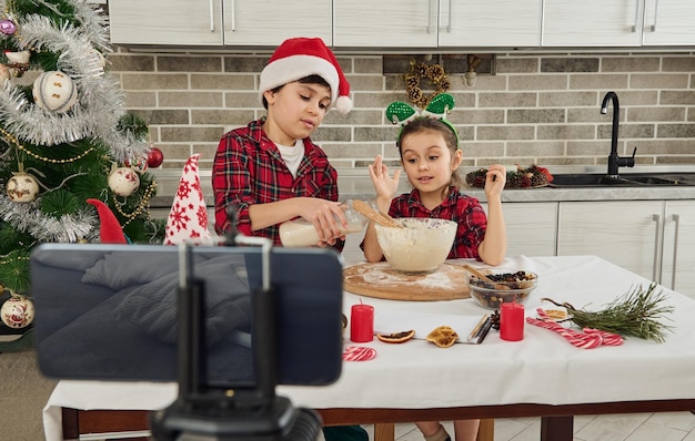 Adorables petits chefs blogueurs pour enfants, un garçon verse du lait dans un bol et une fille pétrit la pâte, enregistrant une vidéo étape par étape d'une recette pour son vlogging sur un smartphone monté sur un trépied. Noël