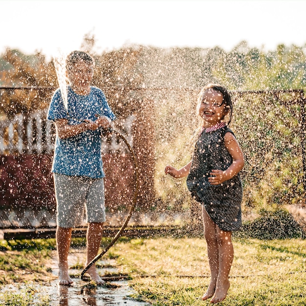 Adorables petits enfants jouant avec un tuyau d'arrosage par une chaude et ensoleillée journée d'été au coucher du soleil