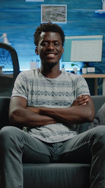 Photo adulte afro-américain assis sur un canapé et regardant la caméra. portrait d'une personne noire aux mains croisées souriant tout en étant à la maison avec la technologie. homme d'origine africaine