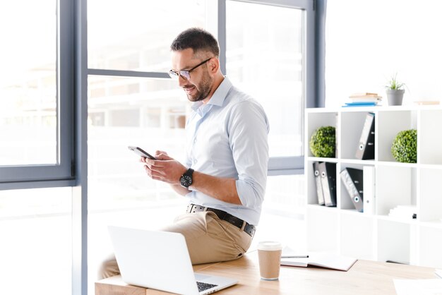 Adulte bel homme en tenue de soirée assis sur la table au bureau près de la grande fenêtre, et tenant le smartphone pour le travail