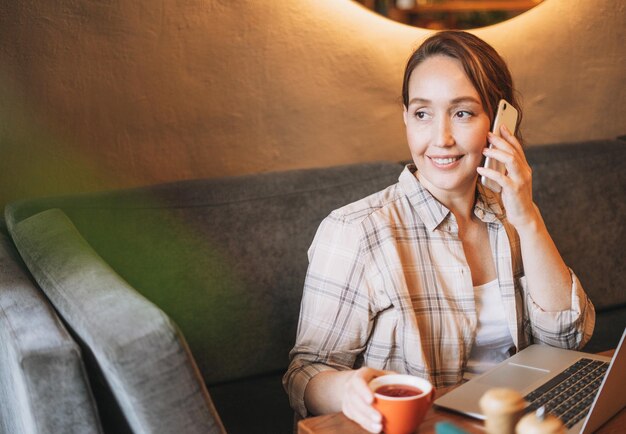 Adulte charmante femme brune en chemise à carreaux travaillant avec un ordinateur portable à l'aide d'un téléphone portable au café