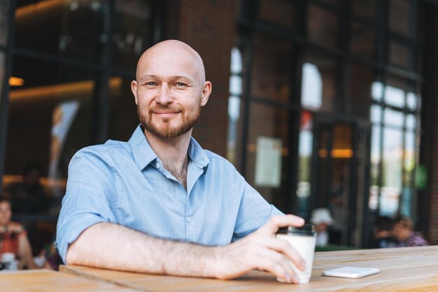 Adulte chauve souriant séduisant homme quarante ans avec barbe en chemise bleue homme d'affaires utilisant un téléphone portable avec une tasse de café en papier au café
