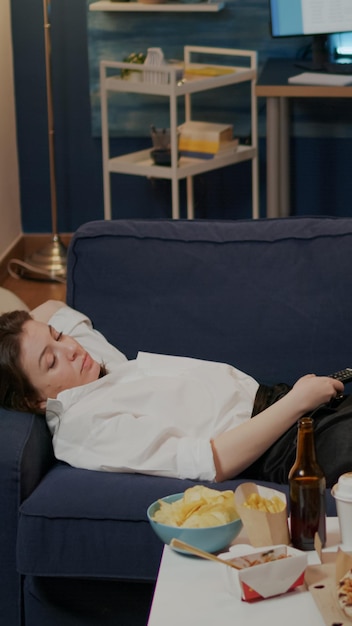 Adulte endormi se reposant sur un canapé après avoir mangé un repas à emporter tout en regardant la télévision dans le salon. Femme s'endormant sur un canapé après le travail et un dîner de restauration rapide à la maison après la livraison