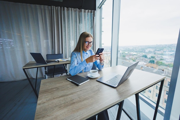 Adulte femme gestionnaire souriante dans des lunettes et une chemise formelle est assise à un bureau et regarde un ordinateur portable Bureau spacieux et lumineux moderne avec une grande fenêtre