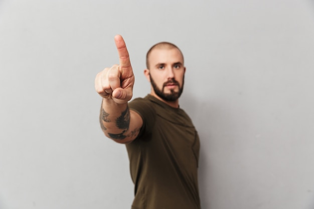 Adulte gars sérieux avec des tatouages sur les mains posant et pointant le doigt vers le haut sur le fond, isolé sur mur gris