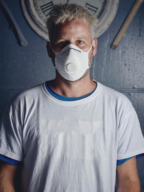 Adulte sérieux aux cheveux blonds, avec des tatouages, portant un masque respiratoire protecteur et un T-shirt blanc, debout près du mur avec une horloge et regardant la caméra.