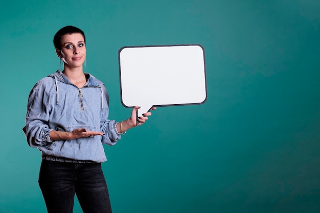 Adulte souriant pointant vers une bulle blanche vide en studio, accessoire d'espace de copie publicitaire pour message texte. Jolie femme confiante regardant la caméra tout en montrant un tableau de modèle vierge