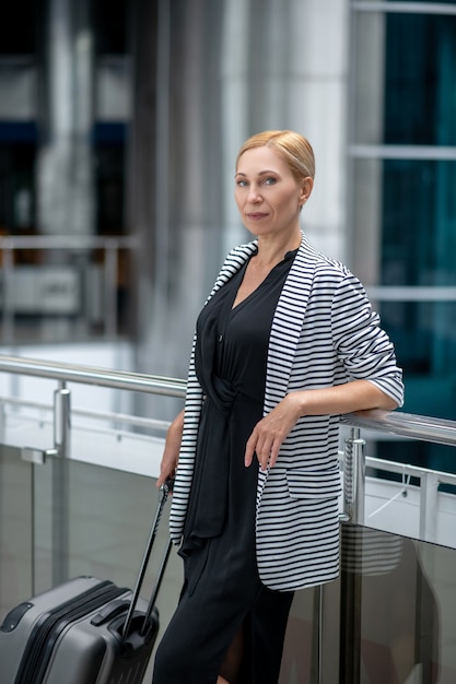 Aéroport, en attente. Femme d'affaires d'âge moyen assez confiante avec une valise debout en attente à l'aéroport dans une humeur positive