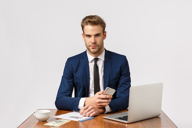 Affaires, entreprise, concept d'entreprise. Patron sérieux assis dans son bureau avec un visage concentré, fronçant les sourcils, tenant un smartphone, attendant un appel important, ayant beaucoup d'argent en jeu