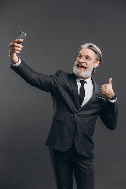 Affaires et homme mature barbu à la mode dans un costume gris faire selfie sur le mur gris.
