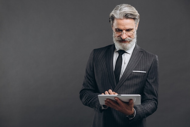 Affaires et homme mature barbu à la mode dans un costume gris faisant des achats en tablette avec sa carte de crédit en or sur le mur gris.