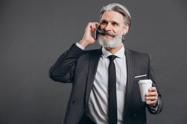 Affaires et homme mature barbu à la mode dans un costume gris parlant par téléphone, buvant du café et souriant sur le mur gris.