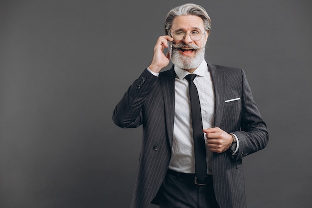 Affaires et homme mature barbu à la mode dans un costume gris souriant et parlant par téléphone sur le mur gris.