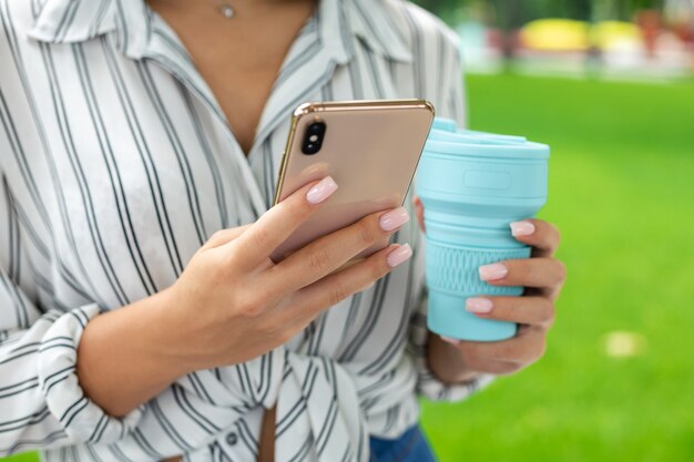 Photo affaires, indépendant, éducation, air frais, technologie et concept internet - une femme blanche vêtue d'une chemise de couleur claire tenant un téléphone et un thermos de café tout en tapant un message.