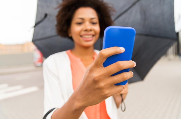 affaires, mauvais temps et personnes et concept - gros plan d'une femme d'affaires afro-américaine heureuse avec parapluie textos sur smartphone dans la rue de la ville