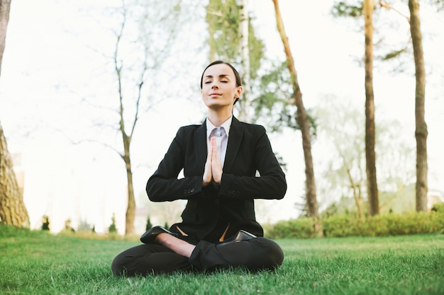 Affaires et méditation Une belle femme d'affaires concentrée dans un costume noir est assise en position de lotus sur l'herbe dans le parc de la ville et se repose après le travail