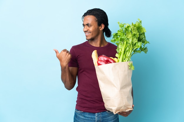African American man holding a épicerie sac isolé sur fond bleu pointant vers le côté pour présenter un produit