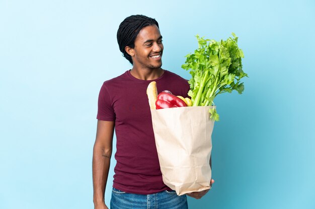 African American man holding un sac d'épicerie isolé sur un mur bleu à la recherche sur le côté et souriant