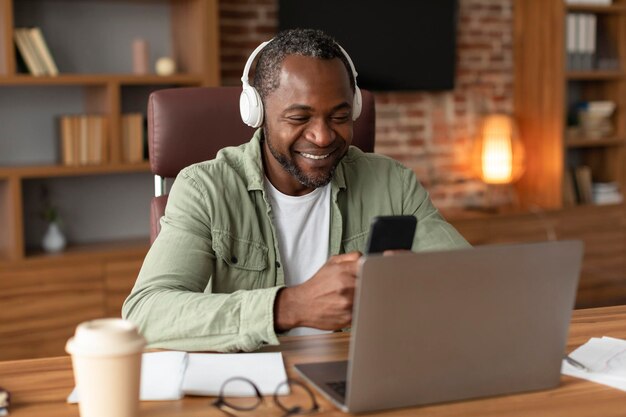 Un afro-américain d'âge moyen souriant dans un casque sans fil tapant sur un smartphone à table avec un ordinateur portable