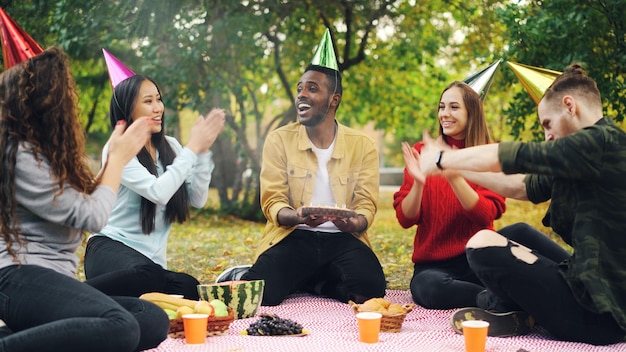 Un afro-américain barbu organise une fête d'anniversaire dans le parc en soufflant des bougies sur un gâteau et en riant en s'étonnant que ses amis applaudissent