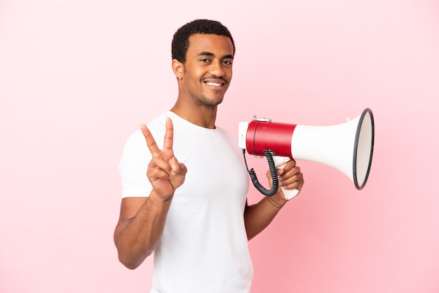 Afro-américain bel homme sur fond rose isolé tenant un mégaphone et souriant et montrant le signe de la victoire